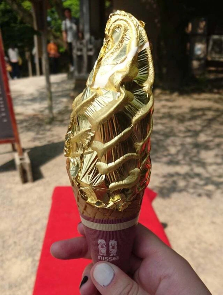 Zmrzlina v kornúte ozdobená zlatou fóliou, držaná v ruke na pozadí exteriéru so slnečným dniom.