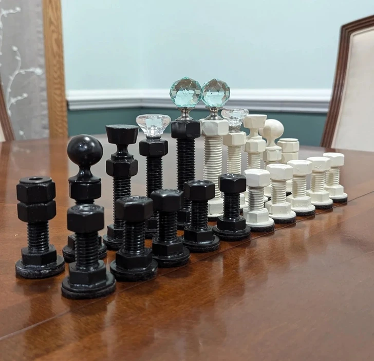 Šachová súprava z matíc a skrutiek, čierne a biele figúrky na drevenom stole, pripravené na hru.