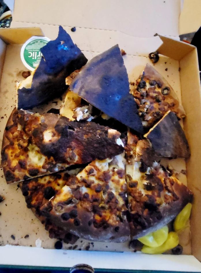 Pizza s prepálenou kôrkou, roztrieštená v škatuľke, doplnená čiernymi olivami a papričkami.