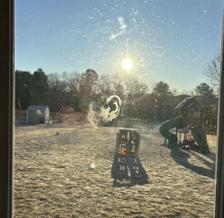 Odraz slnečného svetla na okne so stopou odtlačku ucha, zasnežená záhrada s preliezačkami v pozadí.