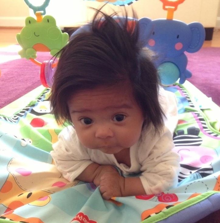 Bábätko na farebnej podložke s hračkami, s roztomilým výrazom a hustými vlasmi, leží na brušku a pozoruje okolie.