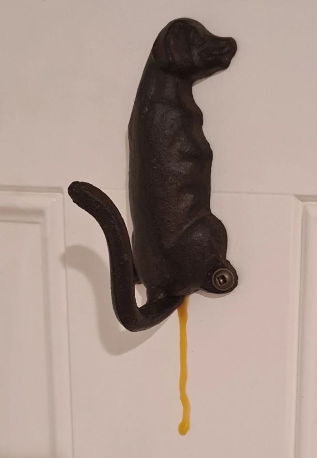 Bronzová figúrka psa s odkvapkávajúcou kvapalinou na bielej stene.