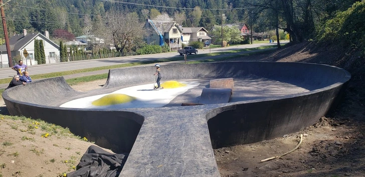 Dieťa jazdí na skateboarde v polokruhovom skateparku na slnečnom dni, v pozadí vidno domy a stromy.