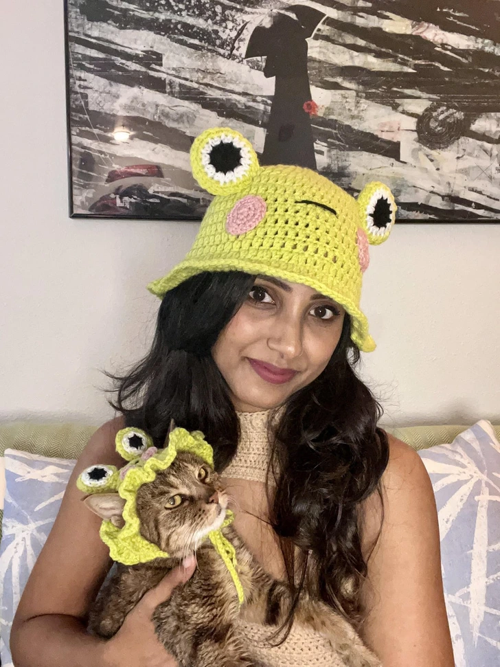 Osoba a mačka v žabích klobúčikoch, sediaci na gauči s umeleckým obrazom v pozadí.