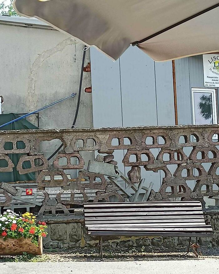 Starý betónový plot plný železa za lavičkou vedľa kvetinovej dekorácie pod slnečníkom.