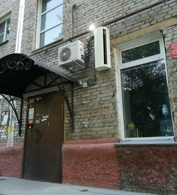 Vstup do budovy s tehlovou stenou, kovovou strieškou a klimatizáciou nad vchodom.