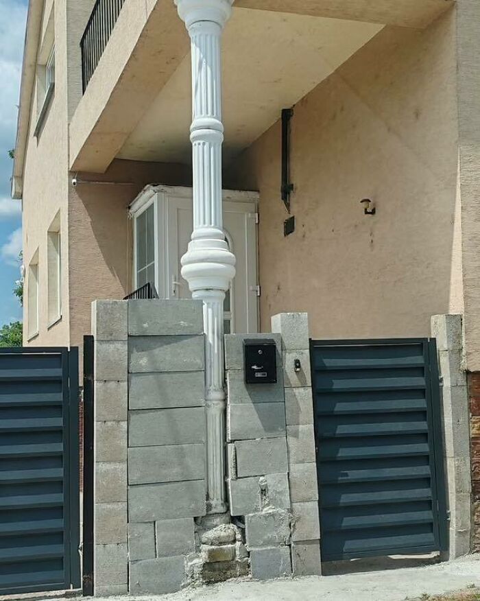 Stĺp v strede dvora vedľa domu s nedokončeným plotom, schránkou a bránou. Neobvyklá architektúra.