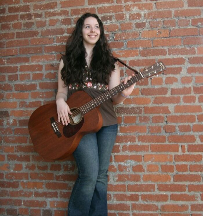 Žena s akustickou gitarou opretá o tehlovú stenu, usmieva sa a nosí džínsy a hnedé tričko.