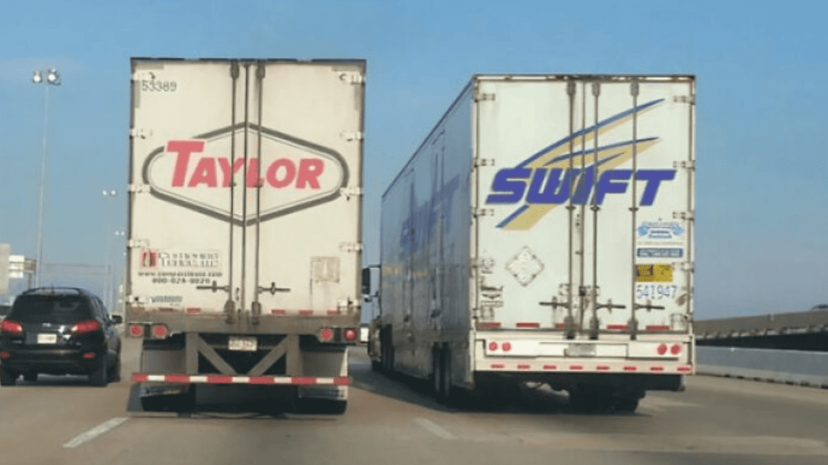 Dve kamiónové nákladné vozidlá na diaľnici, jedno s nápisom Taylor, druhé Swift, vedľa seba na dve jazdné pruhy.