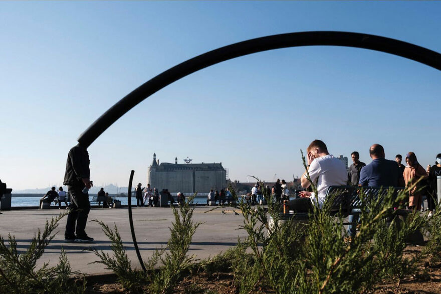 Skupina ľudí odpočíva v parku pri vode, s budovou v pozadí za jasného dňa.