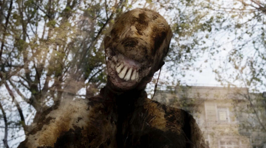 Neznáma postava s desivým úsmevom za sklom, odraz stromov a budov na pozadí.