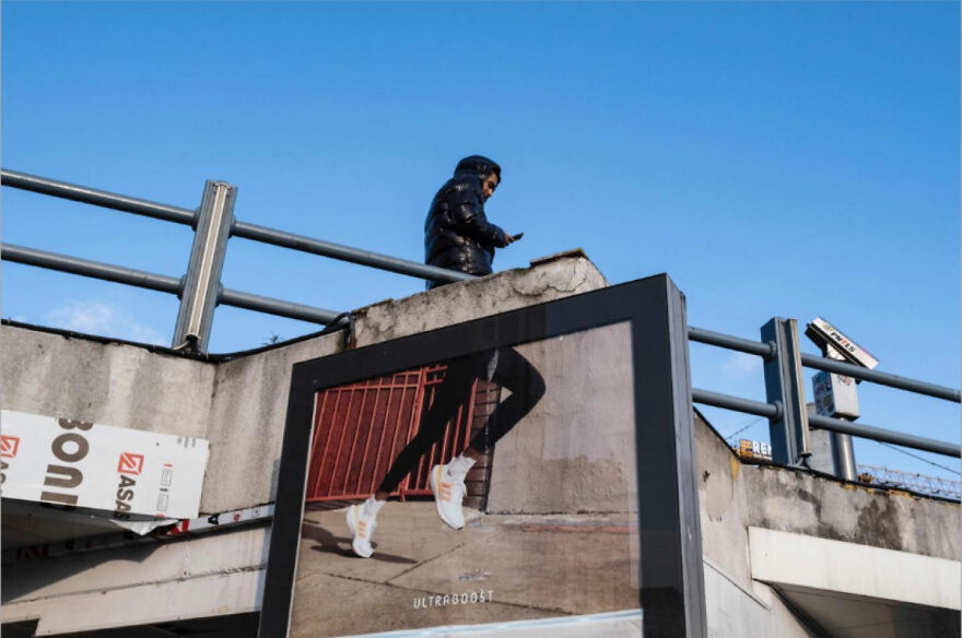 Osoba prechádzajúca cez most nad reklamou na športové topánky, modrá obloha v pozadí.