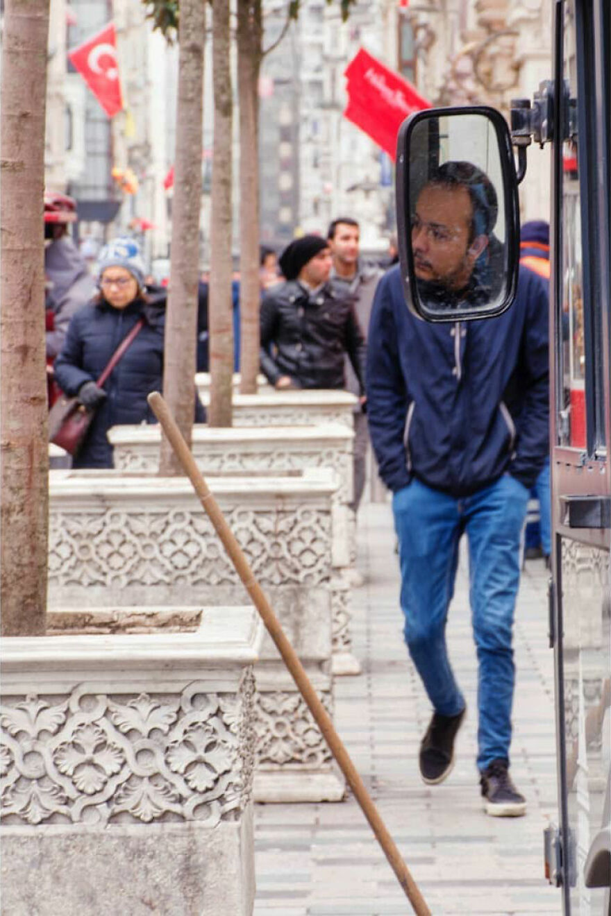 Muž odrazený v zrkadle autobusu kráča po rušnej ulici s vlajkami a kvetináčmi.