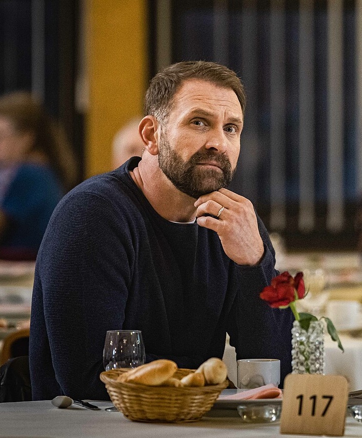 Osoba sedí v reštaurácii pri stole s chlebmi a kvetom, zamyslene sa pozerá do diaľky.