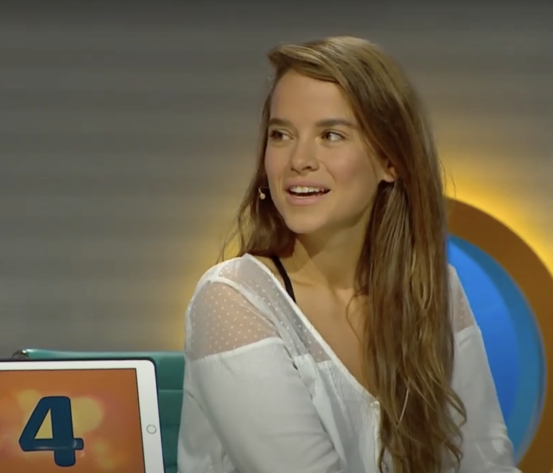 Žena v bielej blúzke sedí v televíznom štúdiu s úsmevom, na stole pred ňou je tablet so zobrazením čísla štyri.