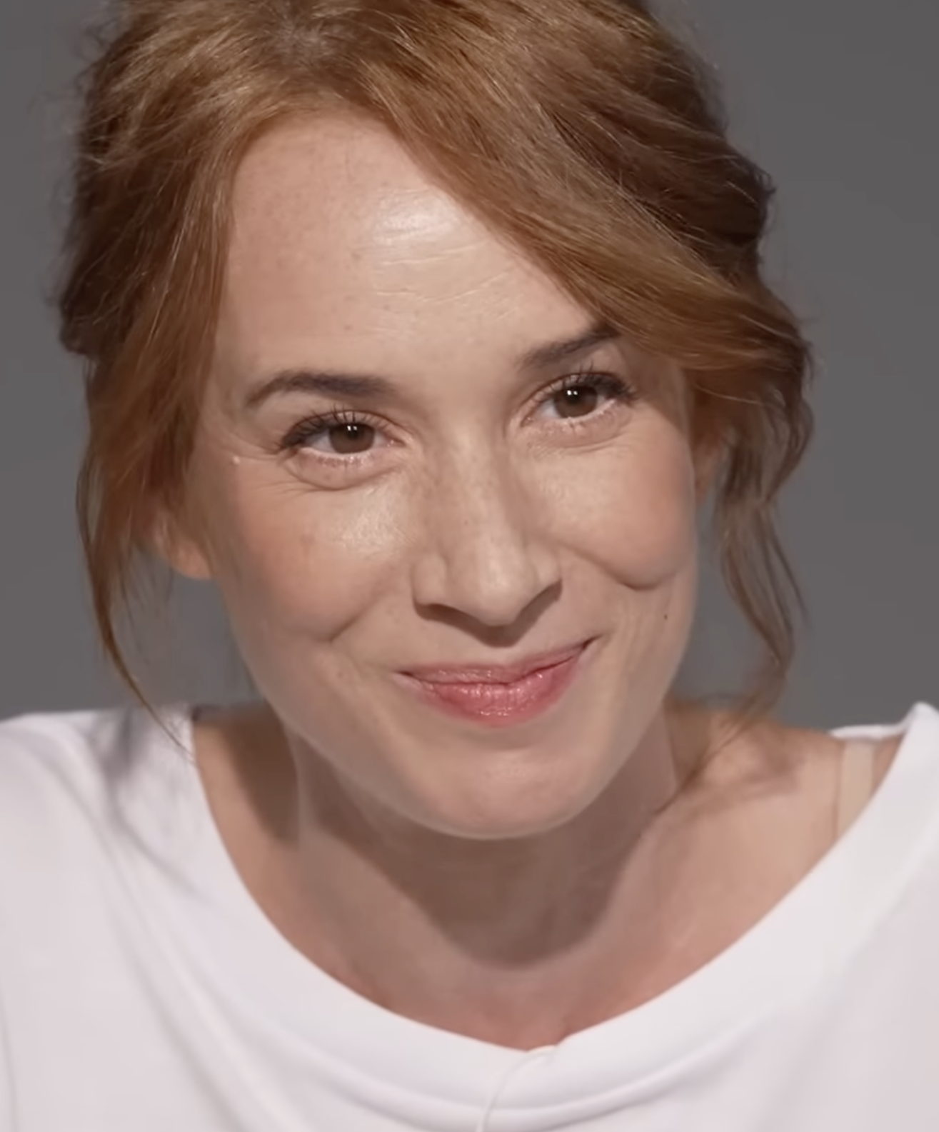 Neznáma žena s červenými vlasmi sa usmieva, oblečená v biele tričko.