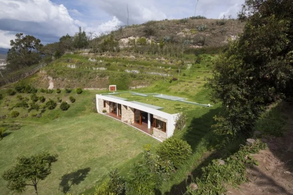 Moderný dom s trávnatou strechou harmonicky zasadený do svahu, obklopený zelenou krajinou.