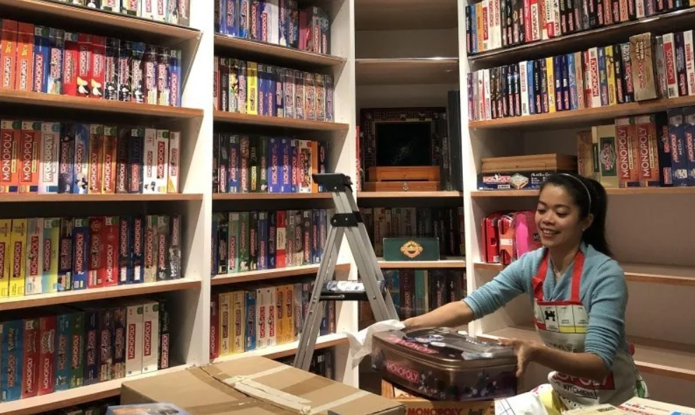 Žena v modrom svetri organizuje zbierku deskových hier na policiach, vrátane rôznych edícií hry Monopoly.