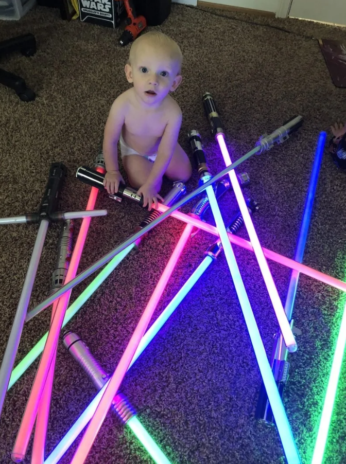 Dieťa sediace medzi rozsvietenými svetelnými mečmi na koberci, vyzerá fascinovane.