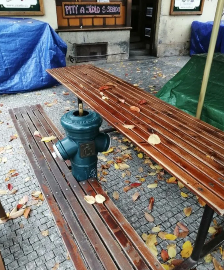 Lavička s hasičským hydrantom uprostred, obklopená opadanými listami na dlažbe pred reštauráciou.