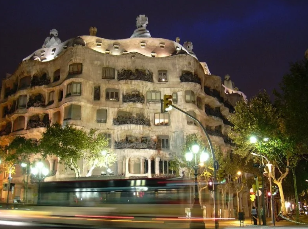 Nočná fotografia ikonickej budovy s vlnitými fasádami, osvetlená pouličnými lampami, obklopená stromami.