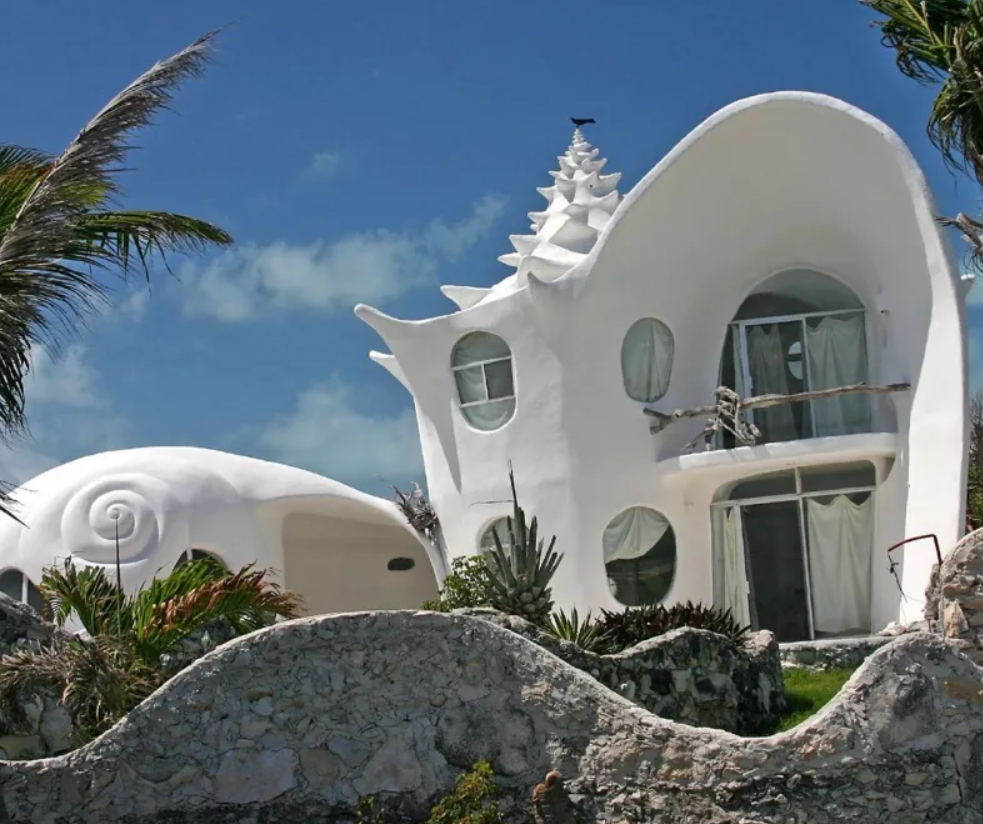 Futuristický dom v tvare ulity s unikátnym dizajnom, obklopený tropickými rastlinami a modrou oblohou.