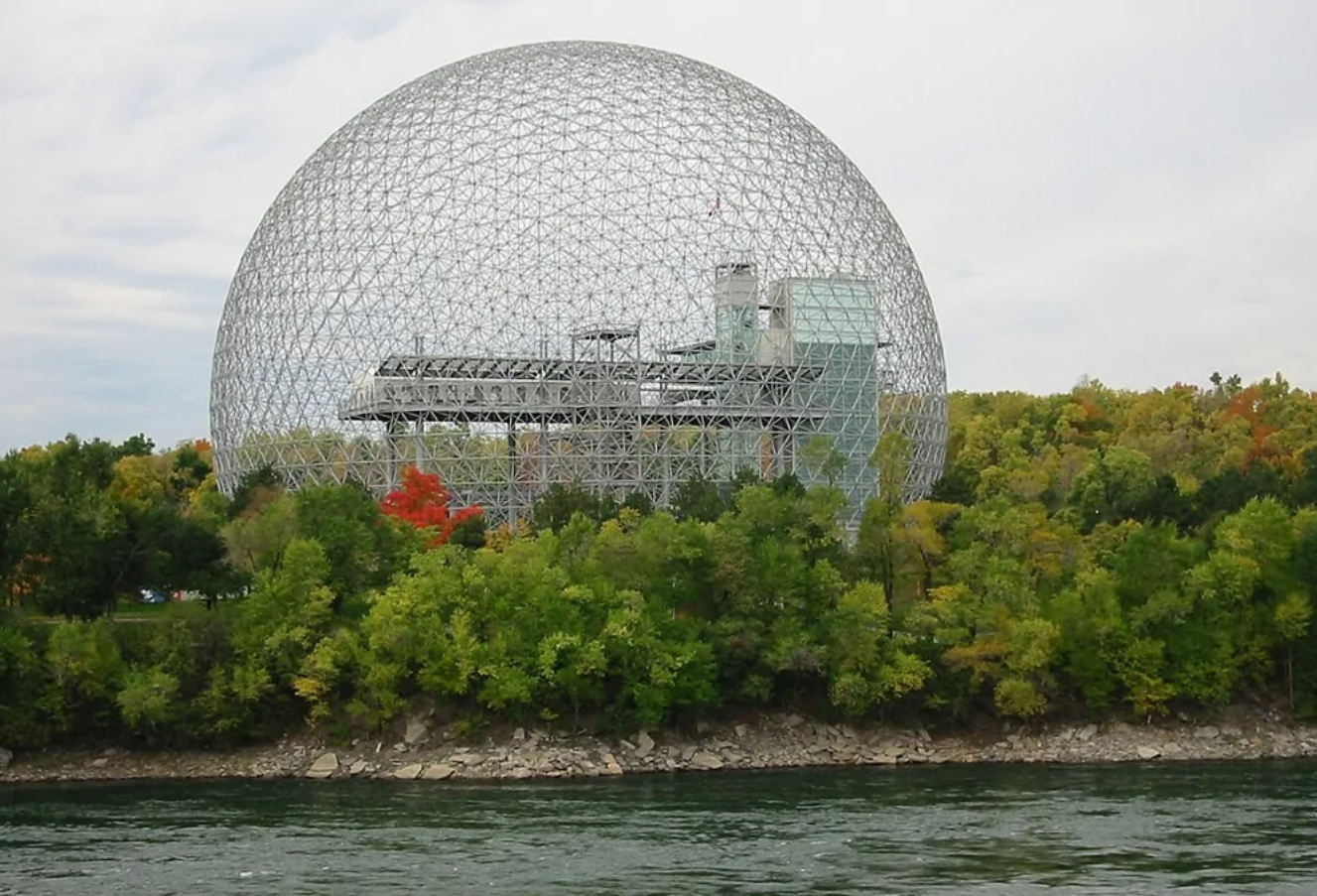 Geodetická kupola uprostred lesa pri vode pod oblačnou oblohou.