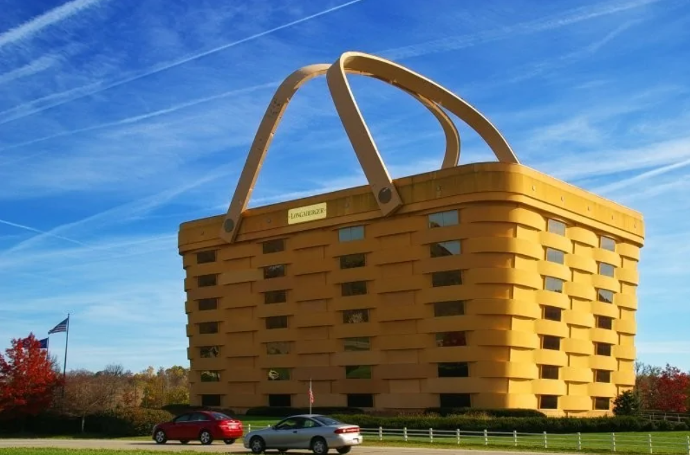 Bežná budova v tvare obrovskej nákupnej tašky pod modrou oblohou, zaparkované autá v popredí.