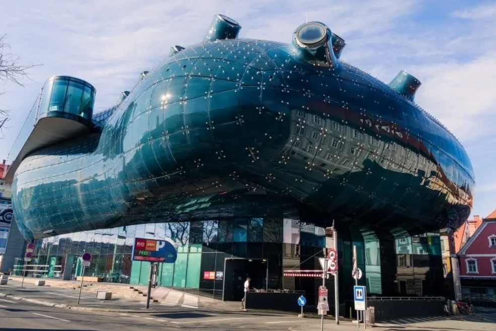 Moderná, futuristická budova s tmavým, lesklým exteriérom v mestskej scenérii, oživená modrým nebom.