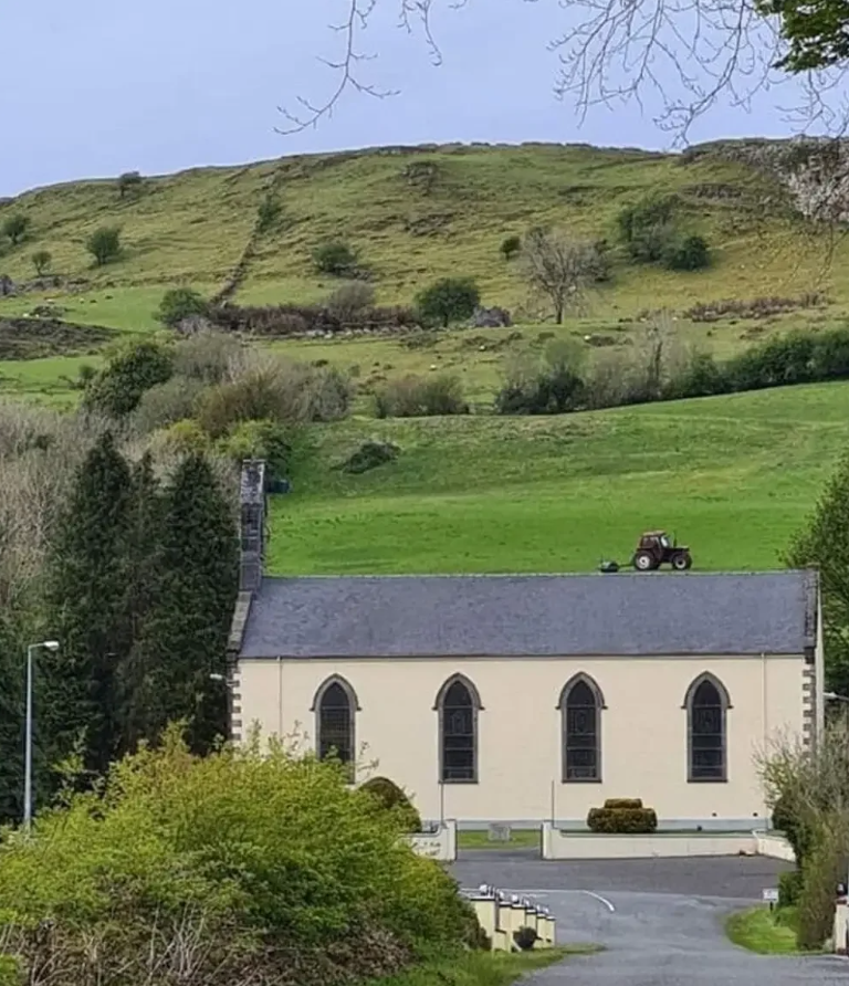 Kostol v malebnej krajine obklopenej zeleňou a kopcami s traktorom v pozadí, všetko pod jasnou oblohou.