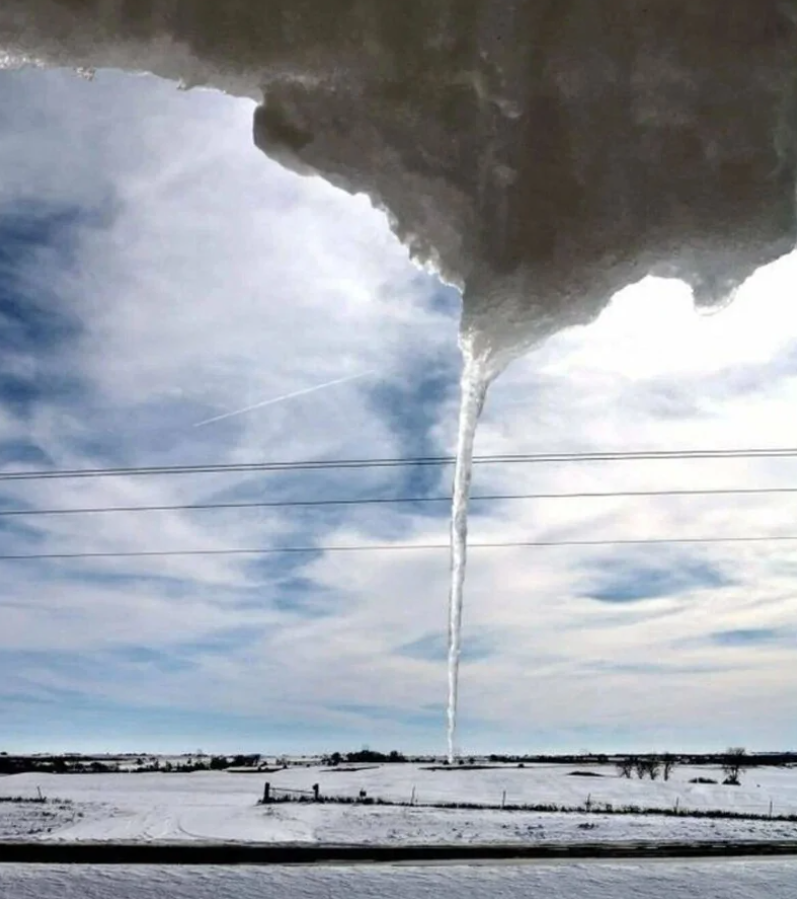 Obrátená krajina s veľkým snehovým cencúľom visiaceho zo strechy, modrá obloha a zasnežené pole v pozadí.