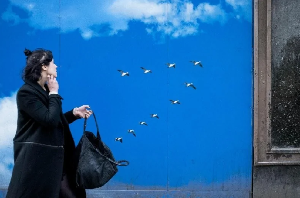 Žena na ulici sleduje múr s modrým nebom a letiacimi vtákmi; kontrast medzi realitou a ilúziou.