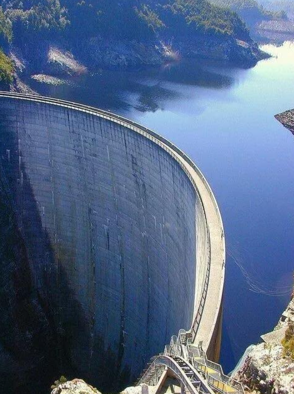 Betónová priehrada obklopená vodou a lesmi pod jasnou modrou oblohou.