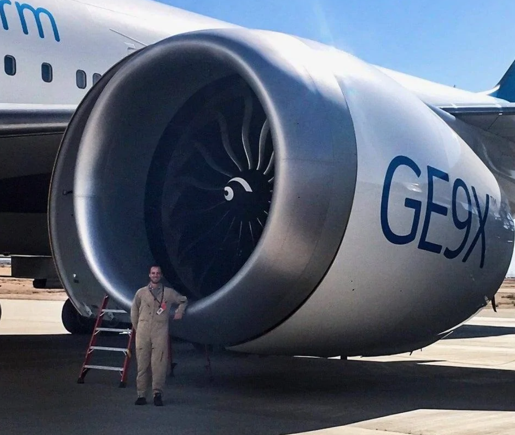 Mechanik stojí pri obrovskom leteckom motore GE9X na letiskovej ploche v jasný deň.