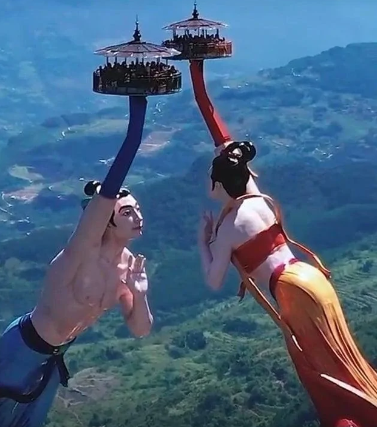 Dve sochy s dlhými ramenami s platformami plnými ľudí vysoko nad malebnou krajinou.