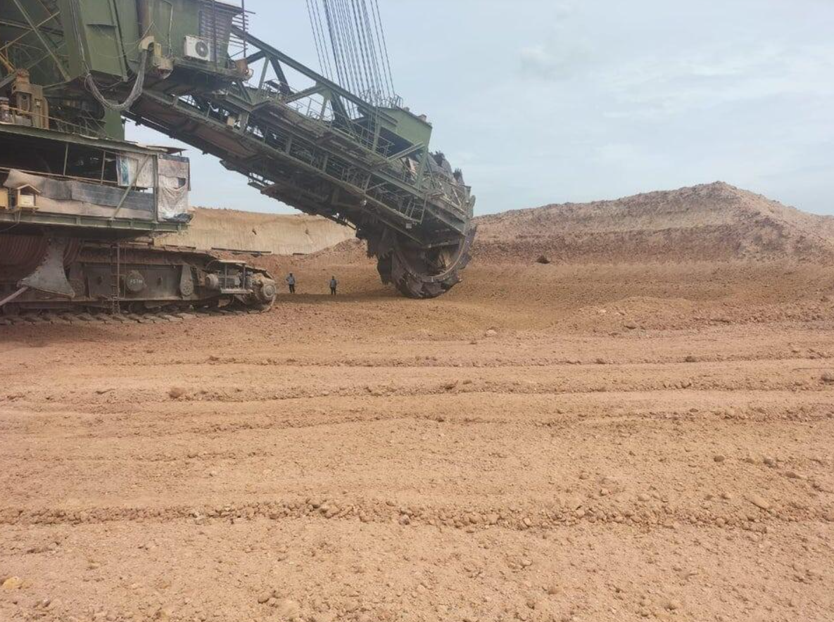 Obrovský ťažobný stroj pracujúci v piesočnej bani, dvoch ľudí sledujúcich pohyb lopatového kolesa.