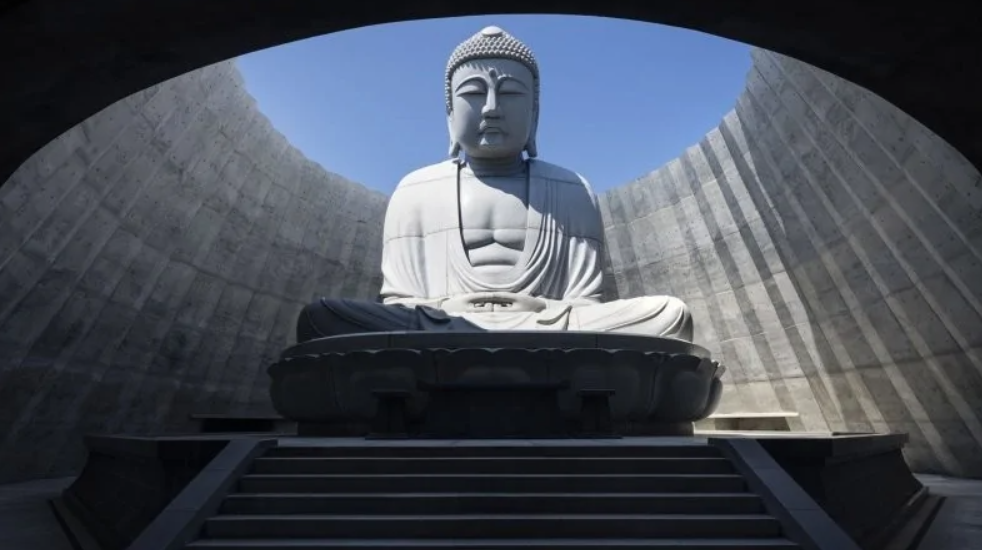 Socha Buddhu v modernom betónovom chráme, pod modrým nebom, obklopená špirálovitými stenami.