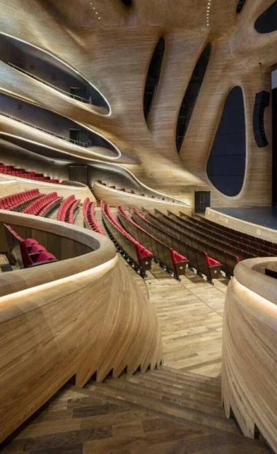 Moderná koncertná sála s drevenými prvkami a červenými sedačkami, zakrivená architektúra interiéru, osvetlené schody.