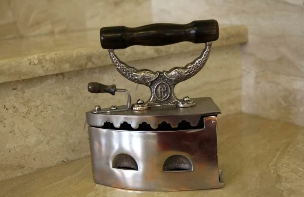 Starodávna kovová žehlička s drevenou rukoväťou, umiestnená na kamennej schode, bez elektrického pripojenia.