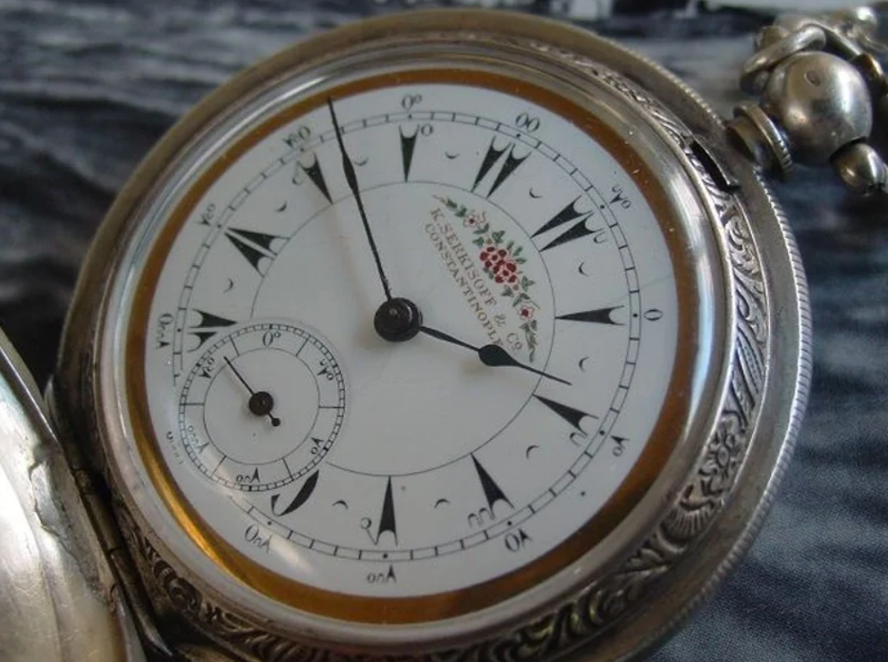 Vreckové hodinky so zdobeným ciferníkom a rímskymi číslicami, jemné detaily gravírovania na kovovom ráme.