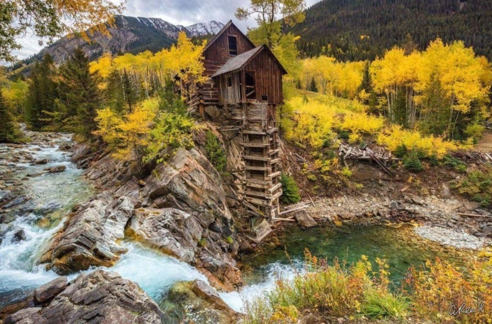Starý drevený mlyn pri horskej riečke obklopený jesennými stromami v malebnej krajine.