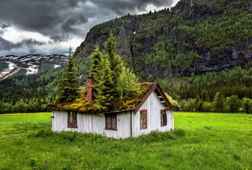 Starý dom s machovou strechou obklopený horami a zelenou lúkou pod zamračenou oblohou.