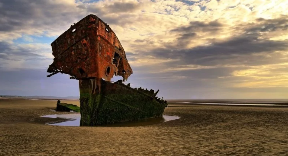 Starý zhrdzavený vrak lode na piesočnatej pláži pri západe slnka, obklopený tajomnou atmosférou.