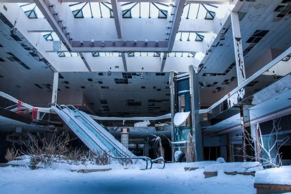 Opustené obchodné centrum pokryté snehom, rozbitý eskalátor a zanedbané rastliny, svetlo prúdi cez strešné okná.