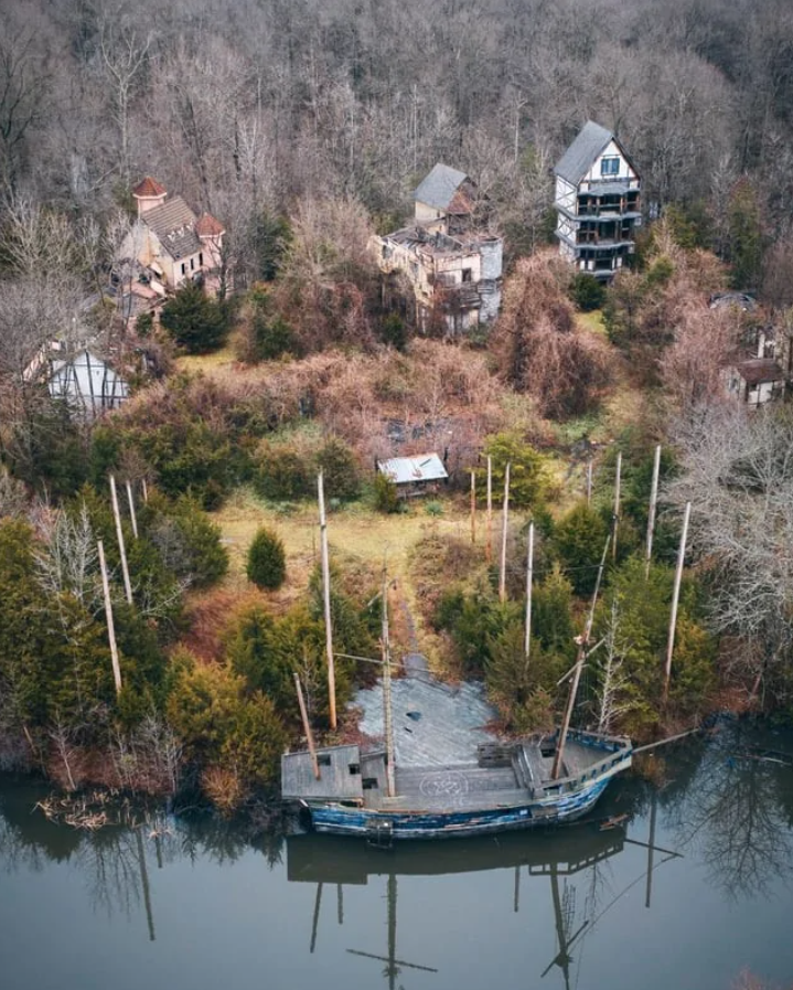 Zarastená opustená krajina s rozpadnutou loďou a starými budovami v hustej lesnatosti.