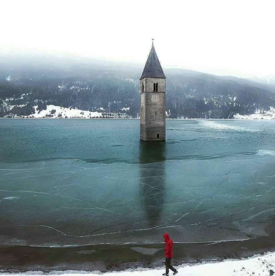 Starobylá zvonica týčiaca sa z zamrznutého jazera Resia v zasneženej horskej krajine.