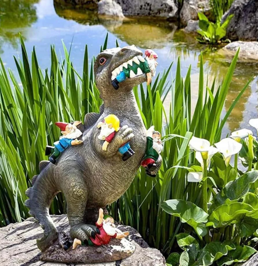 Socha dinosaura a škriatkov pri jazierku obklopenom kvetmi a lístím v záhrade.