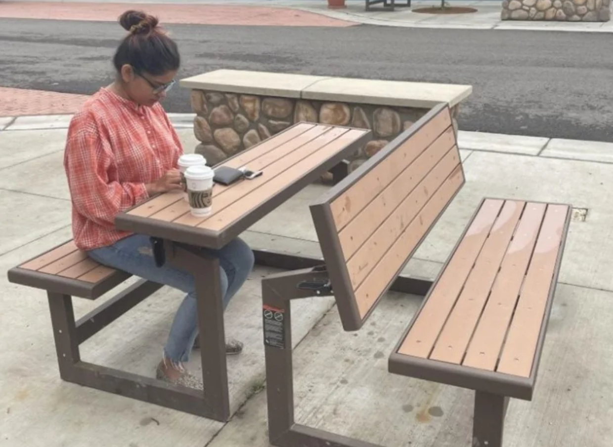 Žena v károvanej košeli sedí pri neobvyklej lavičke vonku s kávou a mobilom na stole.