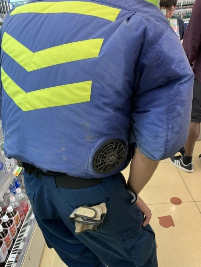 Osoba s veľkým ventilátorom na chrbte v obchode vedľa regálu s nápojmi; pracovné oblečenie s bezpečnostným vzorom.