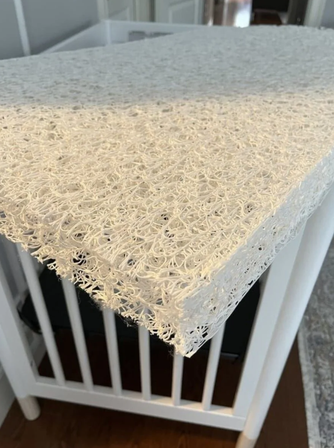 Biela textúrovaná plastová sieťovina na povrchu detskej postieľky nad tmavým matracom.